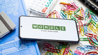 A phone displaying the Wordle logo sitting on a table surrounded by paperclips, pens and notebooks